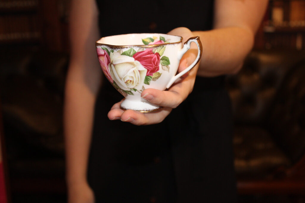 A faded background encompasses a woman's body from her chest to her mid thigh. Wearing a black dress she blends into the background a bit. The center of the frame is her left hand offering a tea cup. The tea cup is white with three roses across the front of it. Her other arm is at her side. She is offering the drink to the viewer. 