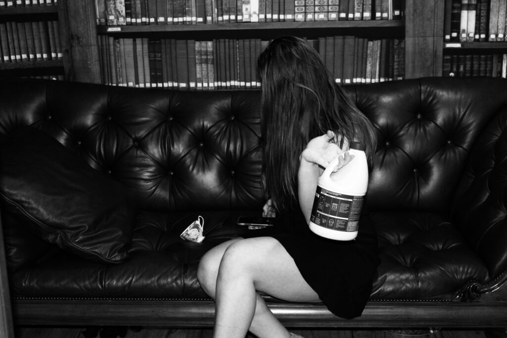 The woman is seated in the same position as the last photo only this time she is facing away towards the towering bookshelves. She has a bottle of bleach tossed over her left shoulder like a hand bag and a tea cup tossed on the couch next to her. 