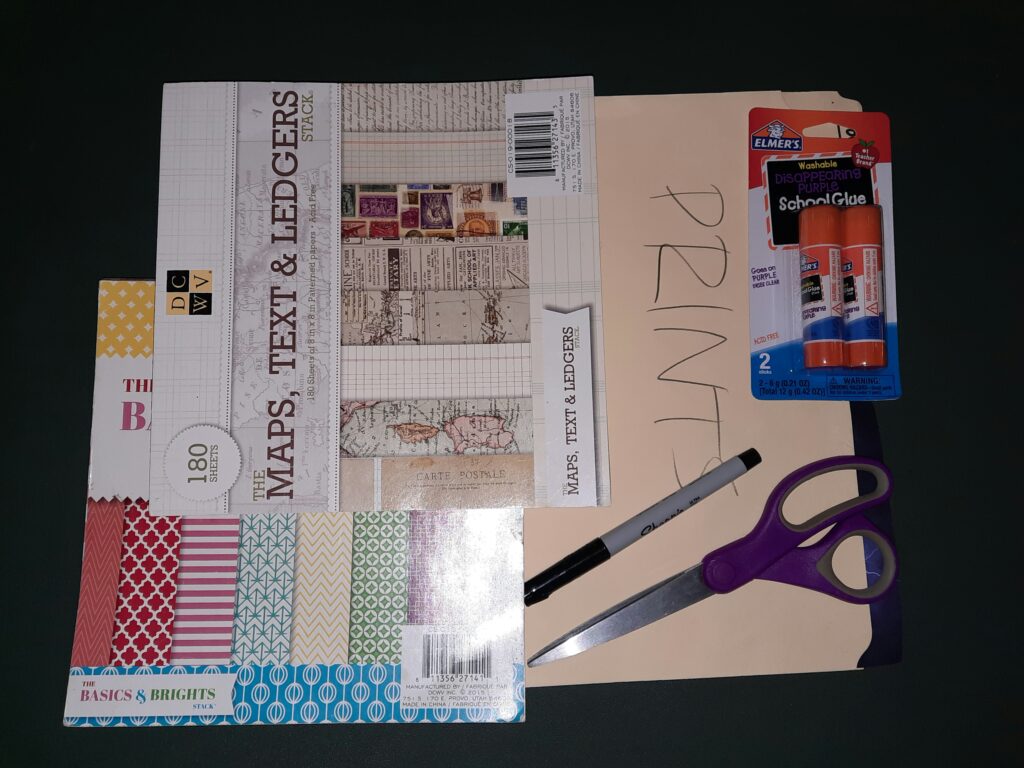 A book with origami pages, a book with scrapbooking texts and maps, a manila folder with “prints” written on it, a pair of scissors, glue sticks, and a Sharpie pen.